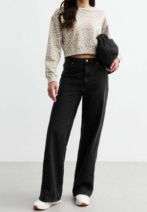 Femme portant un pull court beige à imprimé léopard, un jean noir ample taille haute, des baskets blanches, tenant une doudoune noire.