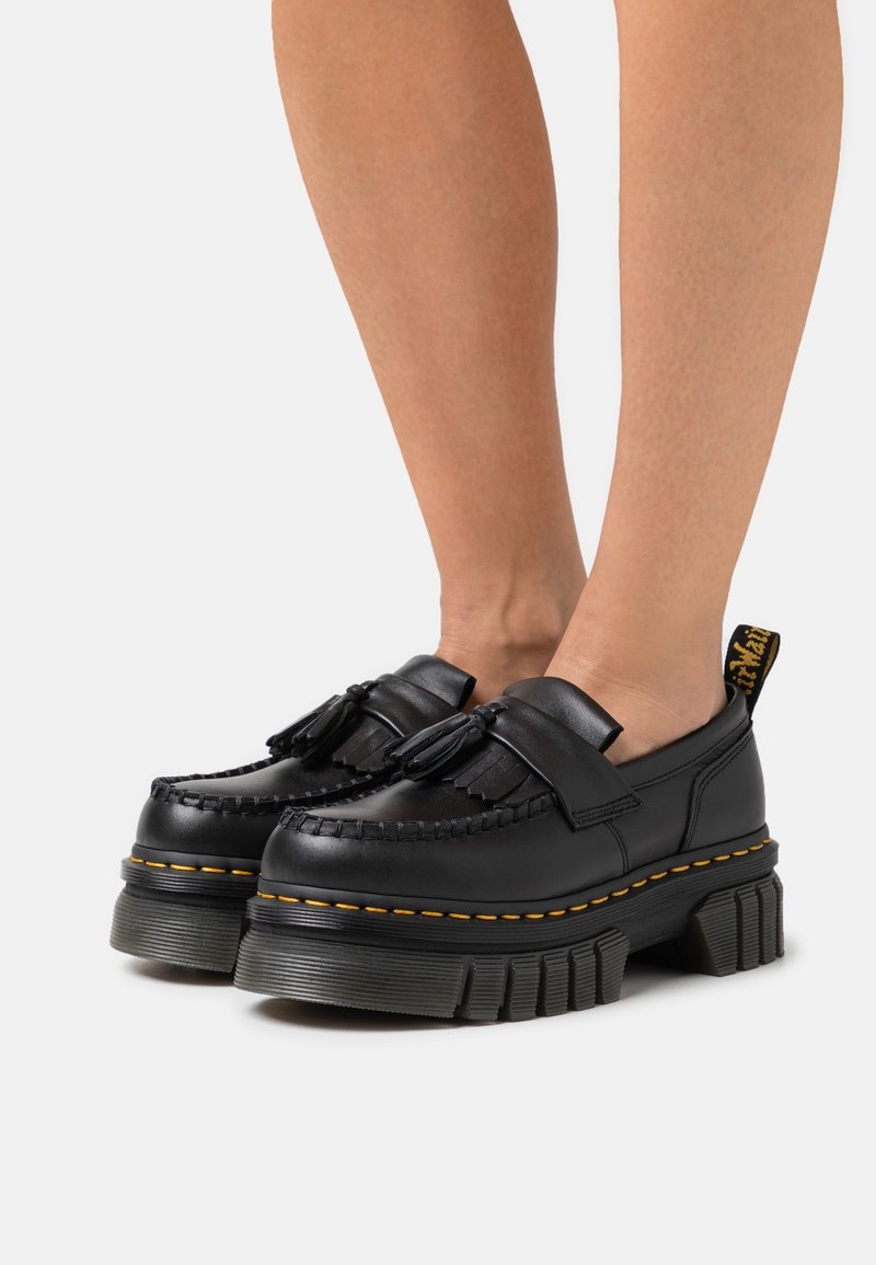 Mocassins en cuir noir avec pampilles et semelles épaisses et robustes portés pieds nus sur un fond uni.