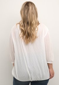 Hvid, gennemsigtig bluse med teksturerede polka prikker, ballonærmer i tre kvarter og en løs pasform, parret med mørkeblå jeans.