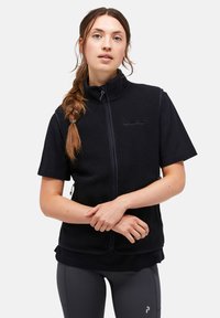 Schwarze Fleece-Weste mit hohem Kragen und Reißverschluss, über einem schwarzen kurzärmeligen Shirt getragen, kombiniert mit engen, dunklen Leggings.