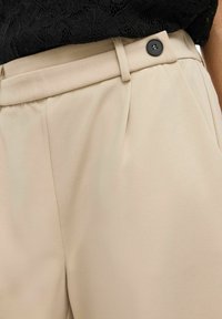 Pantaloni beige a vita alta con chiusura a bottone nera, indossati con un top in pizzo nero infilato dentro.