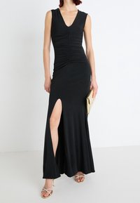 Zwarte mouwloze jurk met V-hals, gerimpeld lijfje, zij-split en vloeiende rok. Paar gouden sandaaltjes met hoge hakken en een gouden clutch.