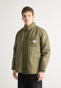Olive green jacket with a collar, zip closure, and two front pockets. Made of sturdy fabric, featuring a patch label on the chest.