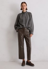 Grijs gestreepte blouse met knopen en ruches op de kraag, bruine leren wijde broek en donkergroene instapschoenen. Matte texturen overal.