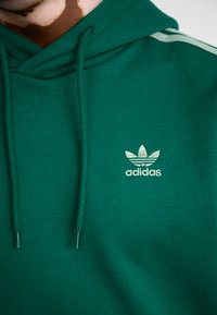Camisola verde com capuz, feita de mistura de algodão, apresentando o logótipo branco da Adidas, cordões e uma superfície texturizada com riscas laterais.