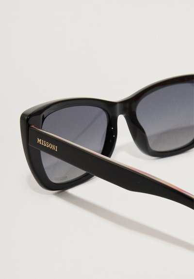 Črne sončna očala z sivimi lečami in drznim, angularnim okvirjem. Na strani se ponaša z zlatim logotipom "MISSONI". Gladka plastična tekstura.