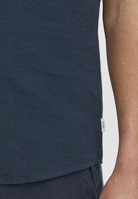 Camisa de algodón de color azul marino con un dobladillo recto, detalles de costura visibles y una pequeña etiqueta de marca en el lado inferior. Tejido suave y texturizado.