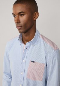 Camicia a righe blu e bianca con accenti rossi sulle spalle e sulla tasca. Presenta una tasca sul petto e una trama di tessuto morbida.