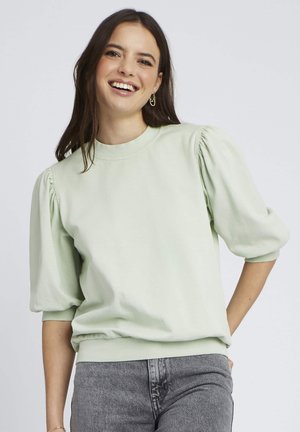 Sweatshirt vert clair avec des manches bouffantes, un décolleté rond, des poignets côtelés et une taille côtelée. Associé à un jean gris. Texture de tissu lisse.