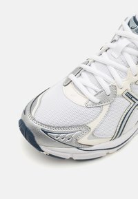 Hvid sportssko med syntetisk mesh-overdel, sølv- og marineblå detaljer, tekstureret tåhætte og tyk gummiydersål. Snørelukning.