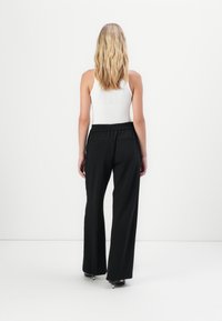 Zwarte wijde broek met een elastische tailleband, met twee achterzakken. Draagt met een witte mouwloze top, gezien van achteren.