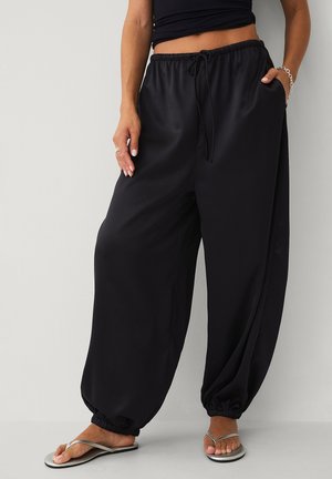 Kvinde iført løse sorte bukser med snøre og elastiske manchetter, en sort crop-top, sølvarmbånd, ringe og sølvfarvede flipflops, stående op ad en almindelig væg.