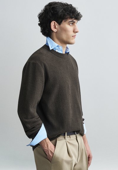 Un pull marron texturé superposé à une chemise à col bleu clair, associé à un pantalon beige plissé. La vue montre un profil latéral.