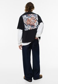 Person med krøllet hår iført lagdelte lange hvide og korte sorte skjorter med abstrakt tryk på ryggen, løse mørke jeans og brune sandaler.