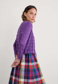 Violet spalvos megztinis su tekstūruotu raštu, derinamas su spalvinga langeline sijonu, kuriame vyrauja raudonos, mėlynos, žalios ir geltonos spalvos.
