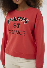 Ženska v rdečem puloverju z napisom "PARIZ 87 FRANCIJA", v kombinaciji z svetlobež hlačami, pred enobarvnim belim ozadjem.