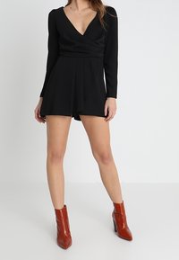 Romper negro de manga larga con un profundo escote en V, pantalones cortos fluidos y una cintura ajustada, combinado con botas de tobillo marrones brillantes.
