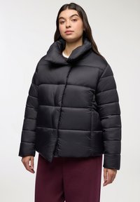 Kobieta w czarnej pikowanej kurtce puchowej z wysokim kołnierzem i burgundowymi spodniami, stojąca na tle jednolitego białego tła.