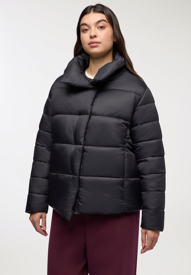 Kobieta w czarnej pikowanej kurtce puchowej z wysokim kołnierzem i burgundowymi spodniami, stojąca na tle jednolitego białego tła.