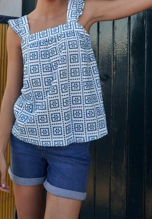 Child wearing blue patterned sleeveless top and rolled-up denim shorts, standing against dark wooden door.