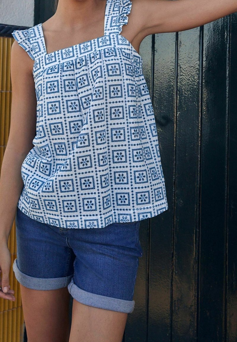 Enfant portant un haut sans manches bleu à motifs et un short en jean retroussé, debout contre une porte en bois sombre.