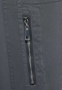 Grått tyg med en mjuk textur; har en silvrig metalldragkedja och en infälld fickdesign. Dragkedjan löper vertikalt längs sömmen.