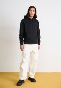 Zwarte katoenen hoodie met trekkoorden en een kangoeroezak; gecombineerd met off-white baggy broek en zwart chunky-soled schoenen op een gele vloer.