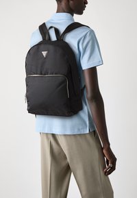 Sac à dos en nylon noir avec des bretelles fines, détail de fermeture éclair argentée, logo triangulaire sur le devant et une texture lisse et futuriste.