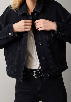 Person, die Knöpfe einer schwarzen Jeansjacke schließt, trägt ein weißes Hemd, das in eine hoch taillierte schwarze Jeans mit Nieten-Gürtel gesteckt ist.