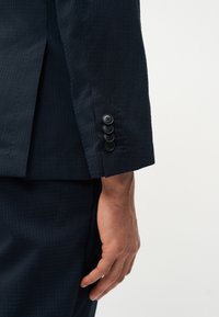 Δεξί χέρι άνδρα που φοράει σκούρο μπλε σακάκι με τέσσερα μαύρα κουμπιά στη μανσέτα, το χέρι χαλαρό δίπλα στο σώμα, σε λευκό φόντο.