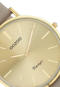 Montre-bracelet en métal doré avec un cadran rond, un fond or clair, des index fins et un bracelet en cuir beige. Présente un texte minimaliste "Vintage".