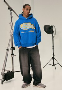 Blå hoodie med gul grafisk text, avslappnade mörka jeans och grå sneakers. Bakgrunden innehåller fotografigrejer.