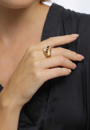 Hand mit nudefarben lackierten Nägeln, die einen Goldring mit bunten Edelsteinen trägt, vor einem schwarzen Kleidungsstück-Hintergrund.