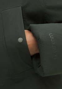 Tmavě zelená bunda s texturovanou tkaninou, viditelným švem, patentním knoflíkem a logem Lululemon na rukávu. Detailní záběr na oblast kapsy.