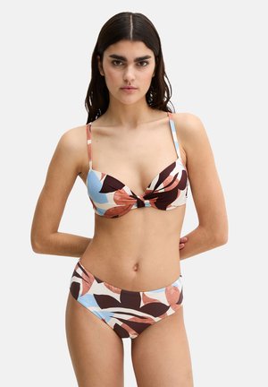 Donna con lunghi capelli scuri che indossa un bikini multicolore con motivo a foglie e spalline sottili, in piedi davanti a uno sfondo chiaro uniforme.