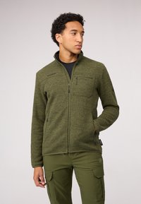 Giacca in pile verde con colletto alto, zip completa, tasca sul petto con zip e dettaglio del logo. Indossata sopra una maglietta nera e abbinata a pantaloni verdi.