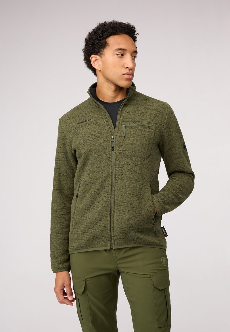 Giacca in pile verde con colletto alto, zip completa, tasca sul petto con zip e dettaglio del logo. Indossata sopra una maglietta nera e abbinata a pantaloni verdi.