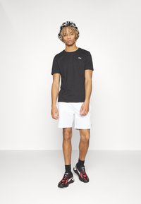 Camiseta negra de manga corta con un pequeño logo, pantalones cortos blancos y zapatillas deportivas negras y rojas. El fondo es blanco liso.