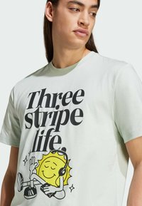 T-shirt in cotone verde chiaro con la scritta "Three stripe life" in grassetto nero, accompagnata da un grafico di un sole giallo con le cuffie.