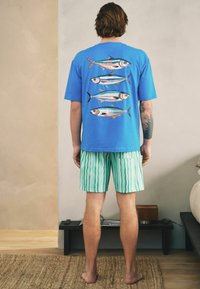Homme debout pieds nus portant un t-shirt bleu avec quatre imprimés de poissons colorés et un short rayé vert dans une pièce minimaliste.
