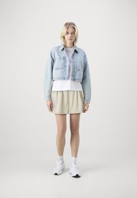 Giacca cropped in denim azzurro chiaro con bottoni frontali e due tasche sul petto, abbinata a una t-shirt bianca e pantaloni corti beige a pieghe. Sneakers bianche.