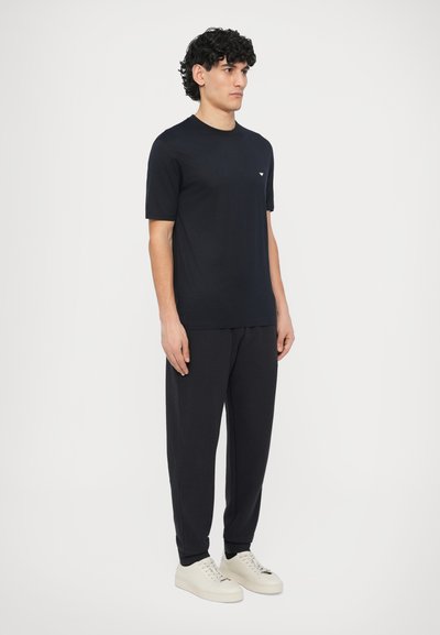 Tmavomodré tričko s krátkym rukávom s nenápadným logom na hrudi, v kombinácii s tmavými štruktúrovanými nohavicami a svetlými teniskami.