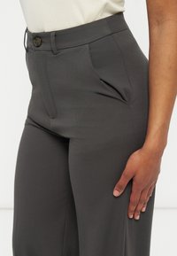 Κοντινό πλάνο ενός ατόμου που φοράει ψηλόμεσα σκούρα γκρι παντελόνια με κουμπί και πλαϊνές τσέπες, το χέρι ακουμπισμένο στον μηρό.