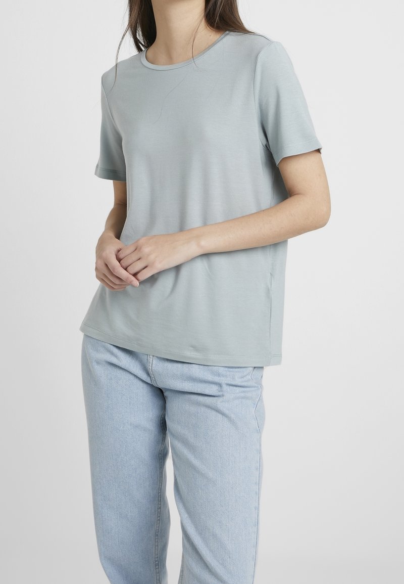 Femme portant un t-shirt bleu clair à manches courtes et un jean bleu clair, debout les mains jointes doucement.