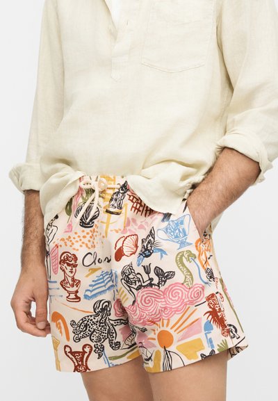 Homme portant un short coloré avec des motifs d'art abstrait associé à une chemise beige à manches longues retroussées.
