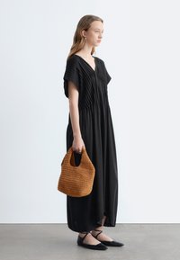 Femme en robe maxi plissée noire tenant un sac à main marron texturé, portant des ballerines noires et des boucles d'oreilles créoles dorées, debout de profil.