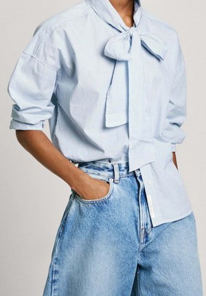 Person trägt eine hellblaue, fein gestreifte Bluse mit einer gebundenen Schleife am Hals und hochgekrempelten Ärmeln, kombiniert mit hellblauen, hoch taillierten Denim-Jeans.
