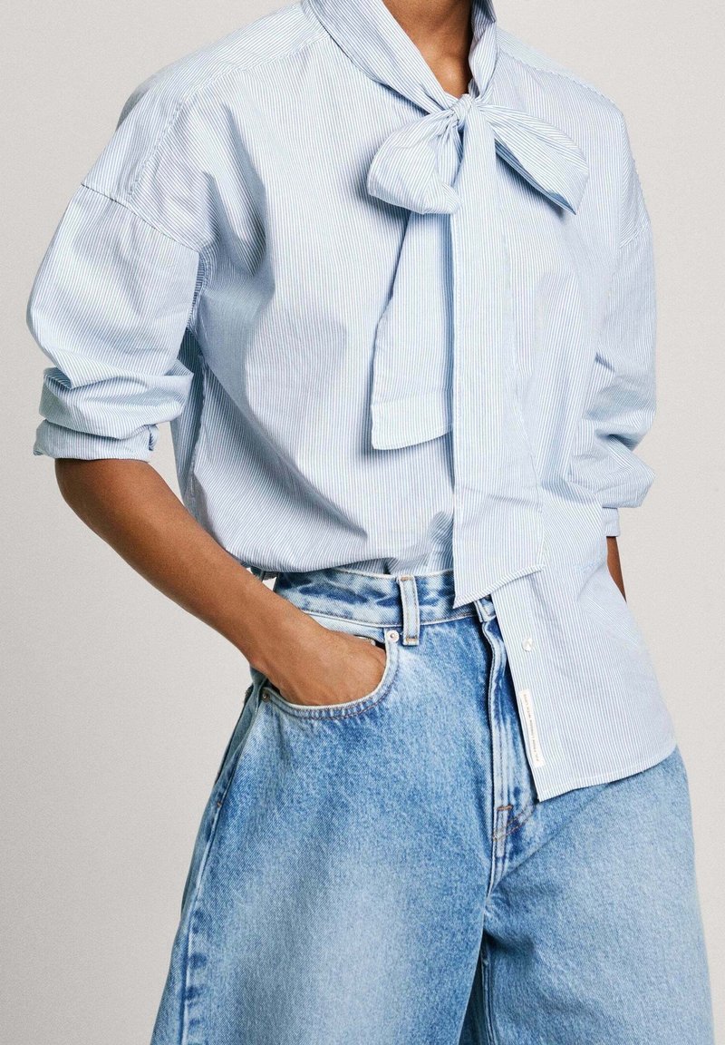 Personne portant un chemisier bleu clair à fines rayures avec un nœud noué au cou et des manches retroussées, associé à un jean en denim bleu clair à taille haute.