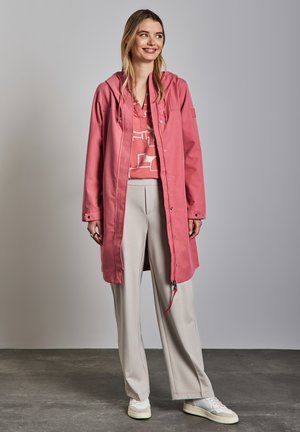 Lyserød vandtæt jakke med hætte, knappakning, sidelommer, parret med lyse bukser og sneakers, sat mod en neutral baggrund.