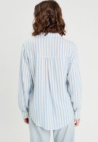 Chemise à manches longues rayée bleu clair et blanche avec une emmanchure arrière, un ourlet arrondi et des poignets boutonnés. Fabriquée dans un matériau doux et texturé.